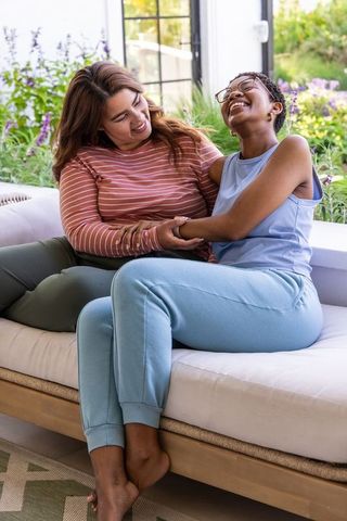 Two women laughing on sunroom bench holding arms, cozy loungewear, friendship moment