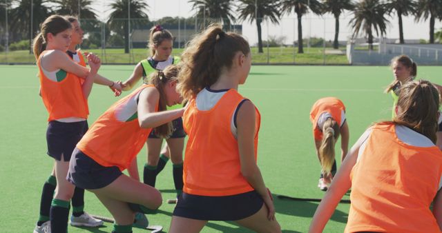 Young female athletes participating in warm-up exercises before starting a field hockey practice session. The girls are wearing orange jerseys and are seen stretching and preparing on a sunny day outdoors. This image is perfect for promoting youth sports, team building, fitness activities, and school sports programs.