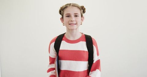 Smiling schoolgirl holding notebook backpack standing against white background