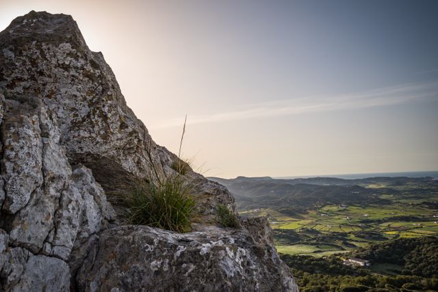 Panoramic view of a rocky cliff overlooking rolling green hills during sunset. The sunlight adds a golden hue to the scenery, promoting a sense of tranquility and serenity. The vivid and natural scenery makes it ideal for use in promotions for outdoor activities, nature retreats, travel advertisements, and inspirational backgrounds.