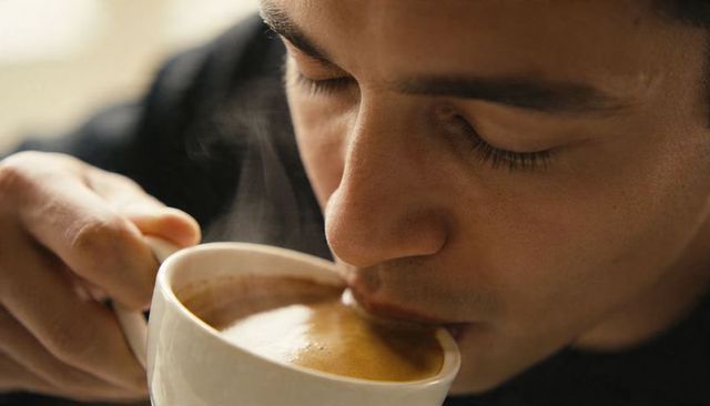 Closeup man savoring espresso crema while holding white mug with steam rising