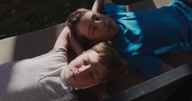 Two young boys relaxing together outdoors on a sunny day, laying on their backs in casual attire. This can be used for themes related to childhood, friendship, summer activities, relaxation, and carefree moments.