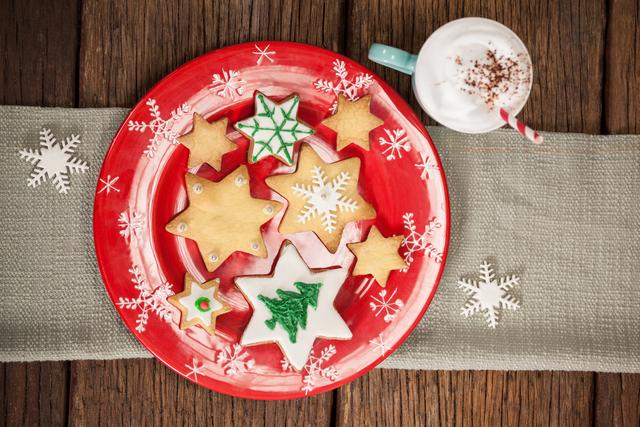 Christmas cookies and a cappuccino arranged on a wooden table create a festive holiday scene. Ideal for holiday recipe blogs, Christmas greeting cards, and social media posts celebrating the festive season. The red plate with star-shaped cookies and the cup of cappuccino add warmth and holiday cheer.