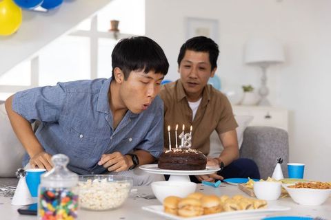 Asian father and son celebrating 18th birthday at home blowing candles on chocolate cake