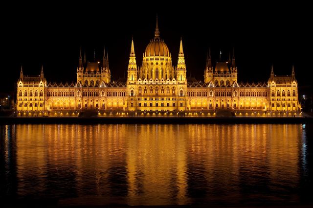 Hungarian Parliament Building glowing beautifully at night, with lights reflecting off the vase Danube River. This iconic landmark in Budapest draws many tourists and showcases the grandeur of European architecture. Ideal for use in travel publications, tourism websites, or architectural magazines.