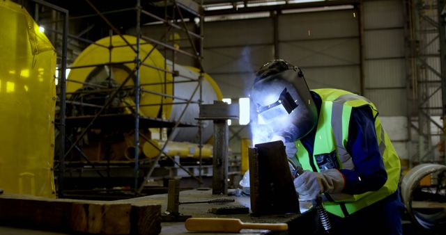 This image of an industrial worker welding metal in a factory is ideal for use in articles about manufacturing, industrial safety, metalworking techniques, and engineering careers. The presence of safety gear like the welding helmet and gloves emphasizes workplace safety. This picture may also be used in brochures or advertisements for industrial safety equipment, vocational training programs, or construction companies.