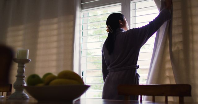Woman in robe opening curtains to let sunlight in at home, concept of starting the day, morning light, relaxation, intimacy, ideal for articles on morning routines, home comfort, lifestyle, wellness.