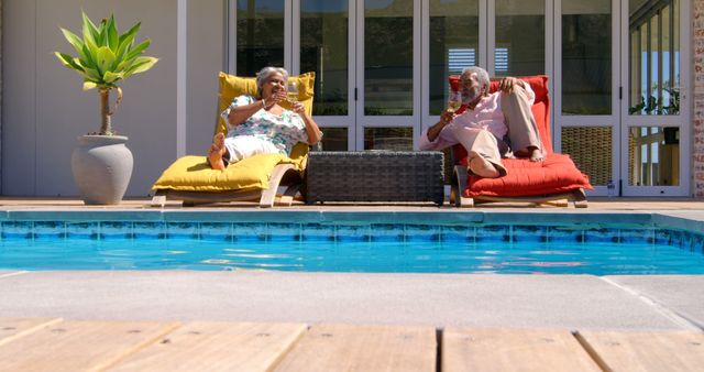 Elderly couple relaxing by the poolside on a sunny day, enjoying drinks while lounging on comfortable chairs. Ideal for use in retirement lifestyle promotions, vacation and leisure marketing materials, health and wellness campaigns, as well as advertisements focusing on senior living and relaxing outdoor activities.