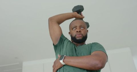 African american man pressing dumbbell overhead in kitchen wearing smartwatch and earbuds
