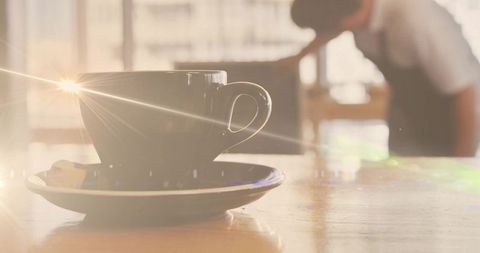 Sunlit coffee cup on saucer catching warm sun flare with biscuit on wooden cafe table