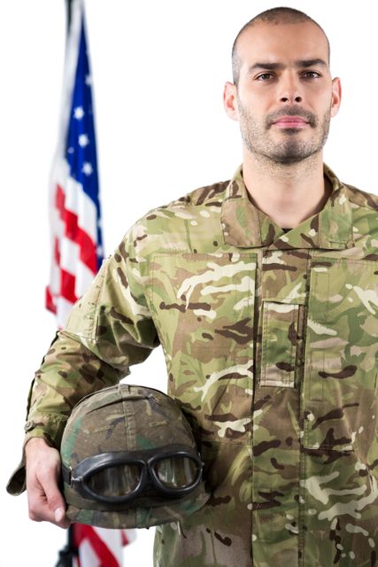This image depicts a soldier in camouflage uniform holding a combat helmet with an American flag in the background. The soldier is standing proudly, symbolizing patriotism and dedication to service. This image can be used for military-related content, patriotic events, recruitment campaigns, and articles about veterans and armed forces.