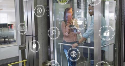Colleagues discussing tablet in glass elevator showing digital business icons and analytics