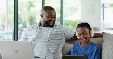 African american father and son relaxing on sofa using laptop and tablet, smiling