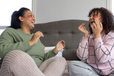 African american friends laughing while applying under-eye gel patches and relaxing on bed