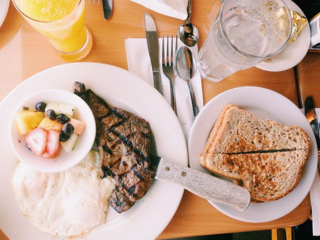 A top-view of a hearty breakfast spread on a wooden table. The meal includes steak and eggs, whole grain toast, a bowl of fresh fruit salad with strawberries, blueberries, cantaloupe, and a glass of orange juice. Perfect for use in food blogs, menus, or culinary magazines to depict a nutritious and delicious breakfast option.