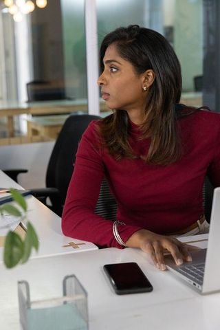 Indian businesswoman working on laptop and checking smartphone in modern open-plan office