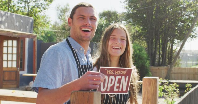 This image shows two cheerful cafe owners holding an 'Open' sign, welcoming customers at the entrance of their outdoor small business. Ideal for promoting small businesses, highlighting entrepreneurship, and customer service themes. Suitable for use in articles, advertisements for cafes, or business-themed promotions.