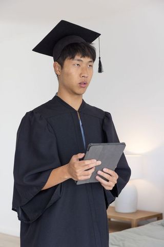 Asian graduate wearing mortarboard and gown holding tablet for virtual graduation ceremony