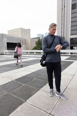 Senior man checking smartwatch after rooftop workout carrying duffel and earbuds