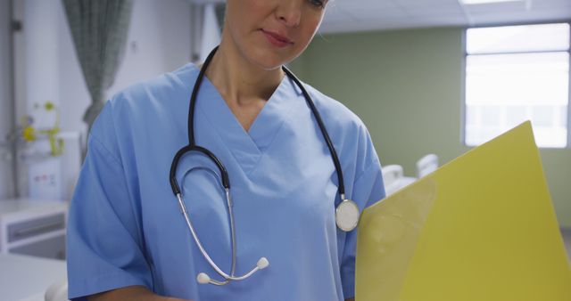 Female nurse is reviewing medical records while standing in a hospital room, wearing a blue uniform with a stethoscope around her neck. This image is suitable for healthcare articles, medical blogs, hospital brochures, and educational materials related to nursing and patient care.