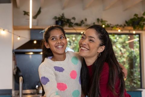 Asian mother hugging smiling daughter in cozy kitchen with polka-dot sweater, warm lights
