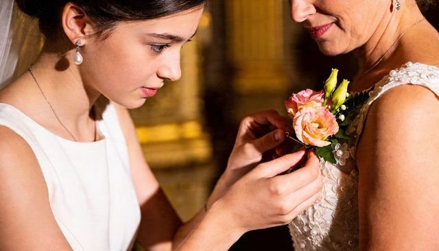 Teenage daughter pinning corsage on mother in lace dress at wedding venue, close-up moment