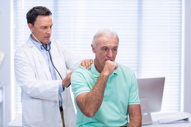 Doctor examining senior patient in clinic. Elderly man in light green shirt coughing while doctor uses stethoscope. Bright, clinical environment with medical equipment in background. Useful for healthcare, medical consultation, senior health, and patient care themes.