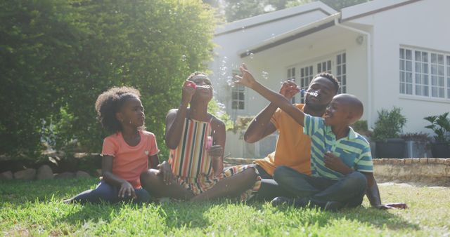 Family sitting on grassy backyard lawn, enjoying summer day while blowing and playing with bubbles. Great for advertising family values, outdoor activities, children’s fun, and summer experiences.