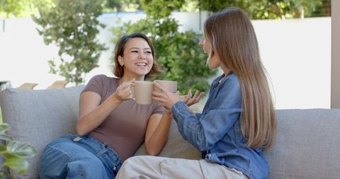 Two women sharing coffee and laughter on light-gray outdoor sofa during sunny backyard chat