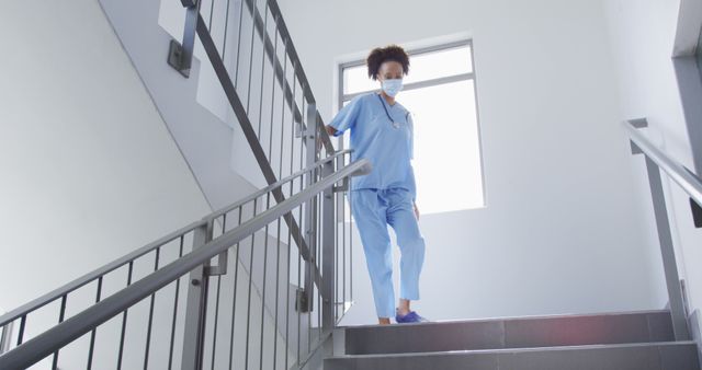 Biracial female doctor wearing face mask running down stairs in hospital. medicine, health and healthcare services during coronavirus covid 19 pandemic.