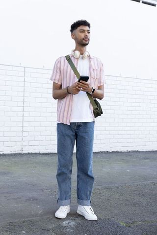 Young man standing with smartphone and headphones against white brick wall, casual urban