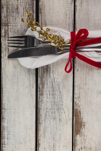 This image features a festive cutlery arrangement with a fork, knife, and spoon tied together with a white napkin and a red ribbon, accented with gold decorations. The rustic wooden table adds a charming, vintage feel. Ideal for use in holiday dining promotions, festive event invitations, or restaurant decor inspiration.