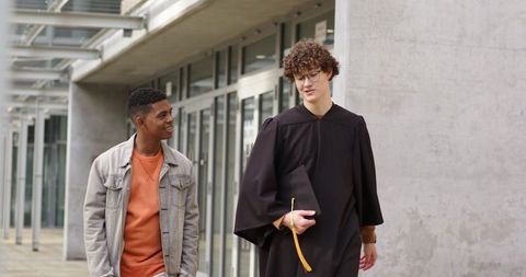 Graduating student wearing black gown walking with friend on modern university campus
