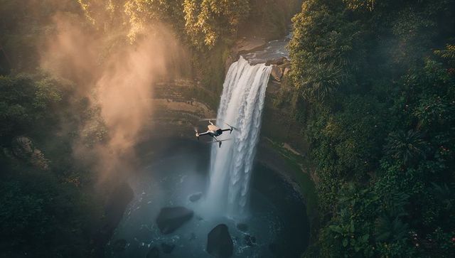 Drone Hovering Over Plunging Waterfall in Tropical Gorge with Mist and Sunbeams