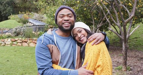 Smiling couple hugging in cozy backyard orchard wearing knit hats and warm sweaters