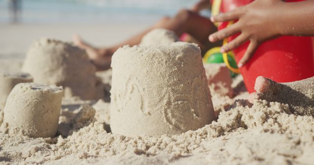 Child building sandcastles with hands and red bucket on a sunny beach. Sand and sea in background. Perfect for themes of summer holidays, outdoor fun, child activities and beach scenes.