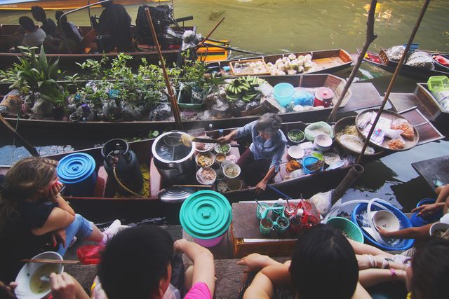 This image shows a busy floating market where a vendor serves customers from a boat filled with various food dishes. The market is vibrant with multiple boats carrying fresh produce, prepared food, and other items. Customers are seated around the water near the vendor boat, engaging in commerce. This image is ideal for use in travel blogs, cultural studies, online articles about street food or markets, and tourism advertisements highlighting unique cultural experiences.