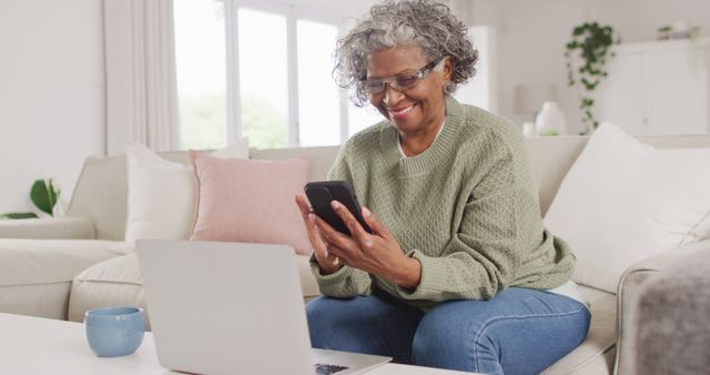 Senior woman sitting on couch using smartphone and laptop, smiling and appears comfortable and relaxed. Ideal for use in advertisements on technology for seniors, lifestyle blogs, and home living concepts.