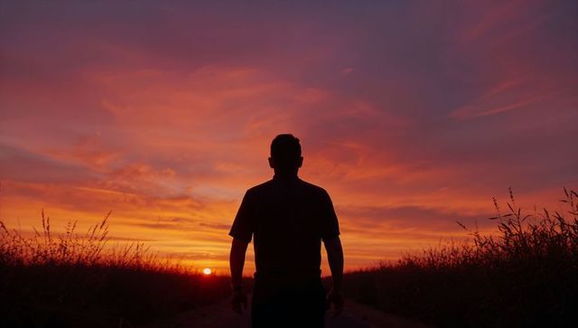 Silhouetted man at sunset on country road watching vibrant orange horizon