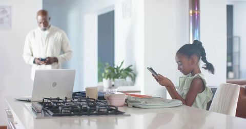 African american father and daughter using smartphones at kitchen island with laptop