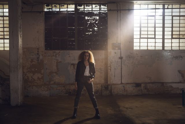 Young Caucasian woman standing confidently in an empty, abandoned warehouse, backlit by sunlight streaming through the windows. She is wearing a leather jacket, exuding a trendy and grunge fashion style. This image can be used for urban fashion campaigns, advertisements for edgy clothing brands, or as a visual representation of confidence and individuality in industrial settings.