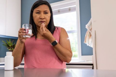 Mature woman taking daily medication with water at kitchen counter wearing smartwatch