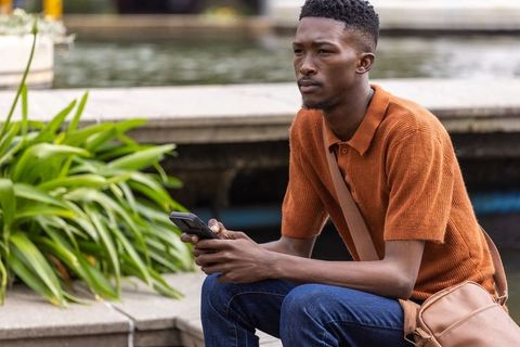 African man sitting on urban steps with smartphone and tan crossbody bag, looking thoughtful