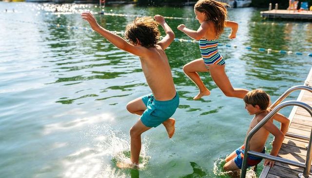 Three kids jumping from dock into sunny lake splashing water in colorful swimsuits
