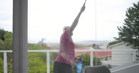 African american man cleaning glass door on balcony with squeegee and blue spray bottle