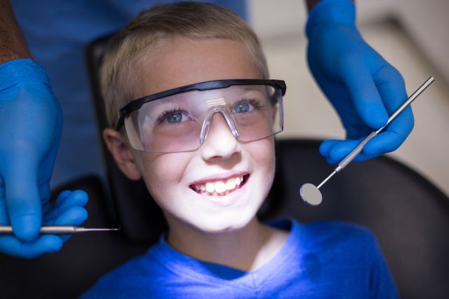Young boy sitting in dental chair smiling while dentist in blue gloves examines his teeth with dental tools. Ideal for use in articles about pediatric dentistry, dental health, oral hygiene tips for children, or promotional materials for dental clinics.