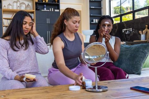 Three african american women sharing skincare routine on sofa using mirror and roller