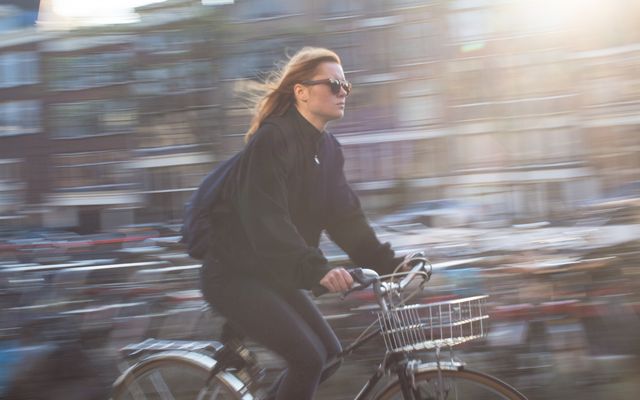 Woman riding a bicycle through an urban environment with motion blur effect. She is wearing sunglasses and casual clothing. Sunlight shines through, enhancing the setting with a dynamic and lively feel. Ideal for use in content related to urban life, transportation, healthy lifestyle, and daily activities.