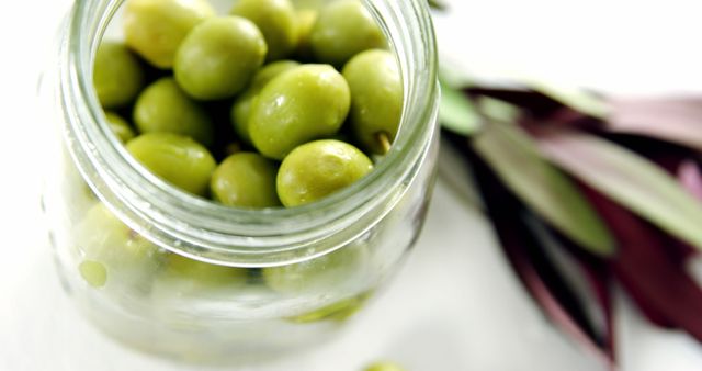 Close-up view of green olives in a glass jar on a white background, perfect for use in food blogs, recipe sites, healthy eating guides, and Mediterranean cuisine features.