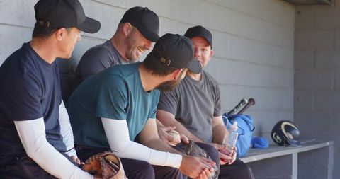 Baseball teammates laughing and talking in dugout while holding gloves and bats